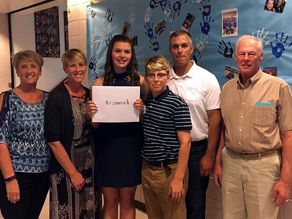 Six people pose for a photo in a hallway, a girl in a navy dress holds a sign that reads 'Krznarich' with a blue handprint mural behind them.