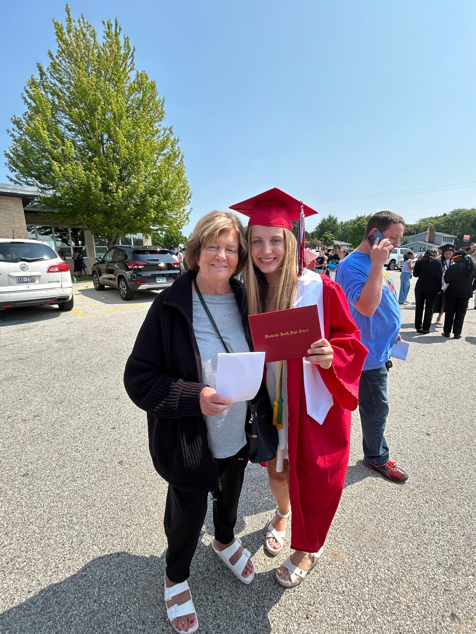 Graduate in red cap and gown holding a diploma, posing with an older woman outdoors in a parking lot.