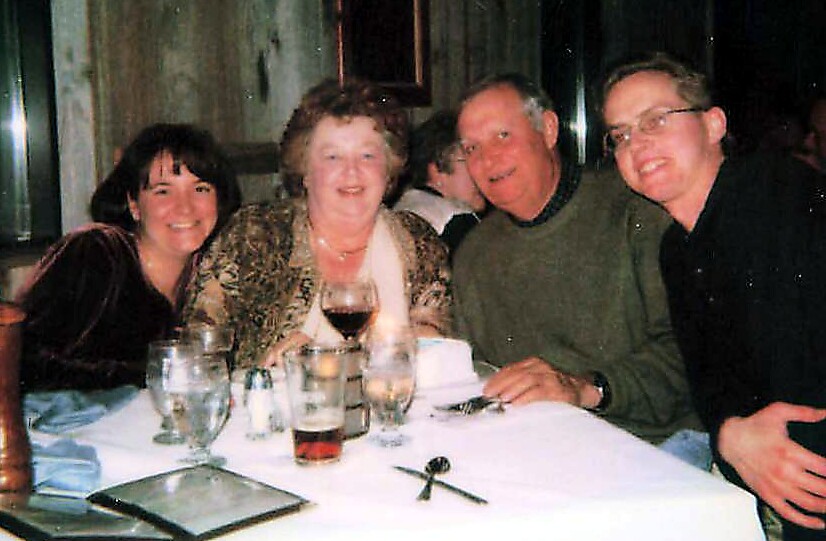 Four friends sit closely around a table in a restaurant, smiling with wine glasses and a pitcher in view.