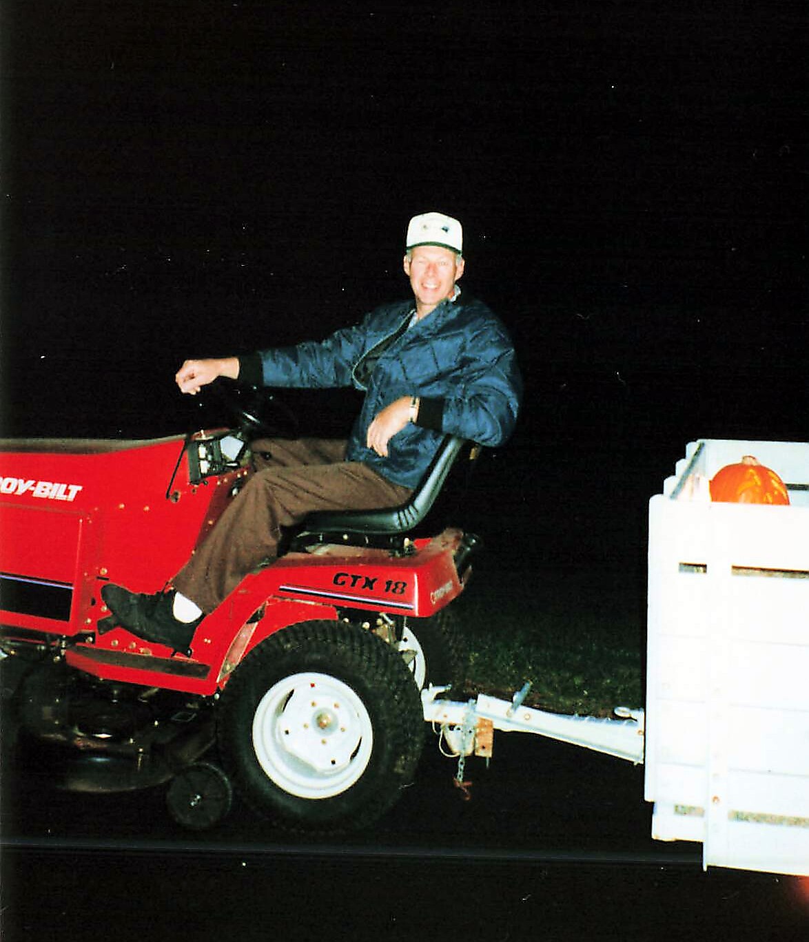 Man in a blue jacket and white cap smiles while seated on a red GTX 18 riding mower attached to a white trailer at night.