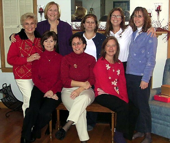 Group of nine women posing together indoors, wearing festive sweaters and smiling for a holiday photo.