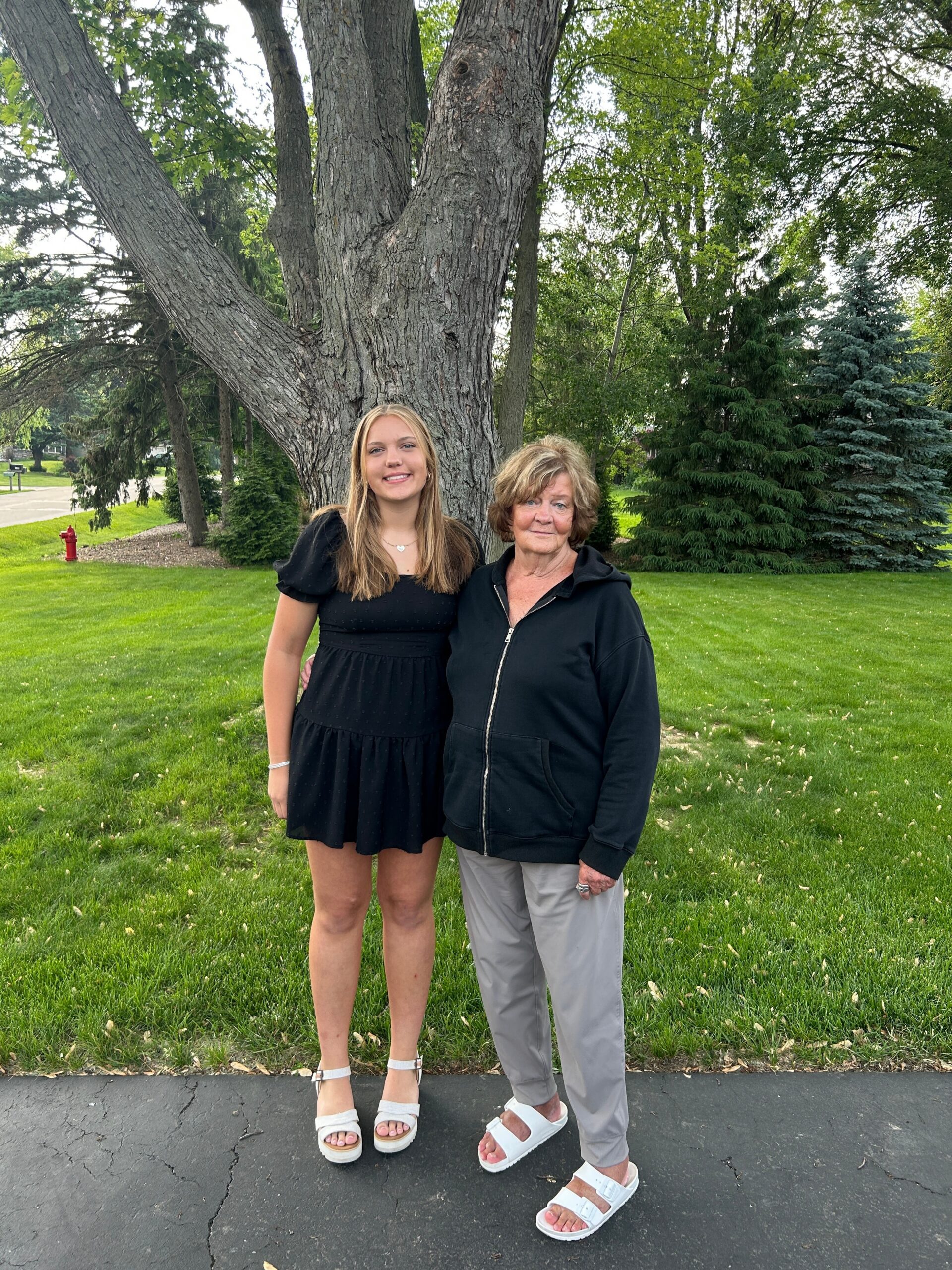 Two women posing together outdoors in a park, standing in front of a large tree on a grassy area by a paved path. First woman in a black dress, second in a black jacket and light pants.