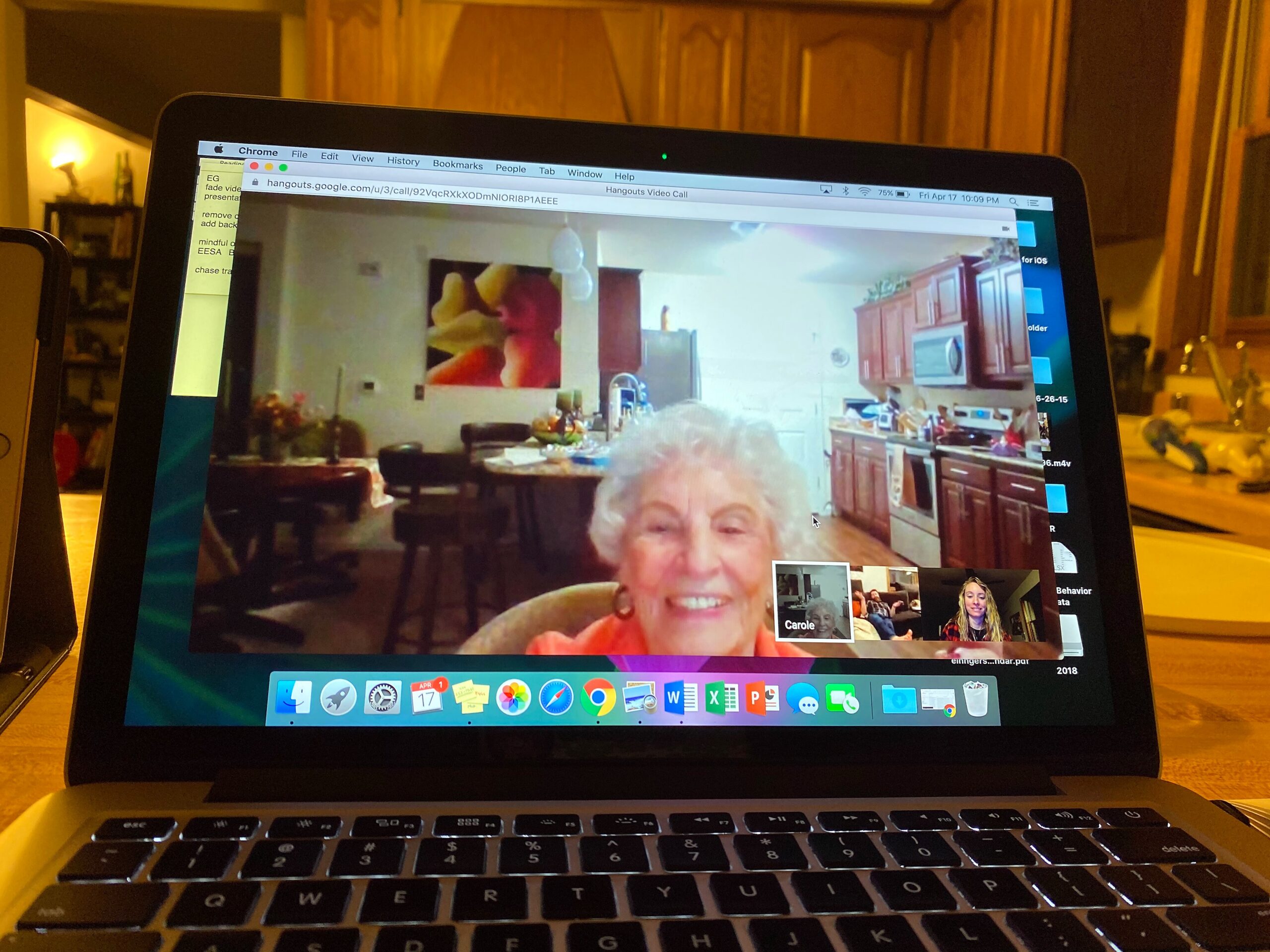 Video call on a laptop screen: smiling older woman in the foreground with a kitchen background and several participants in small video tiles at the bottom.