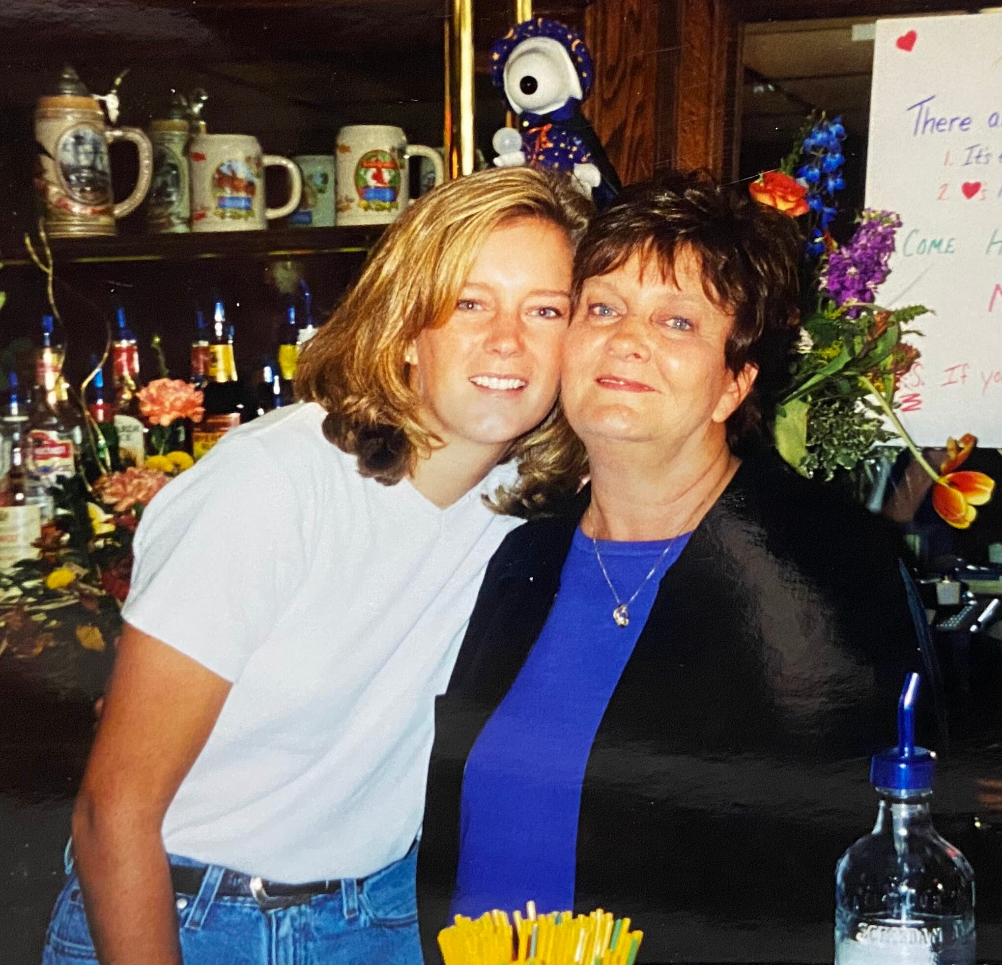 Two women smiling at a bar, with beer mugs on shelves behind them and colorful flowers nearby.