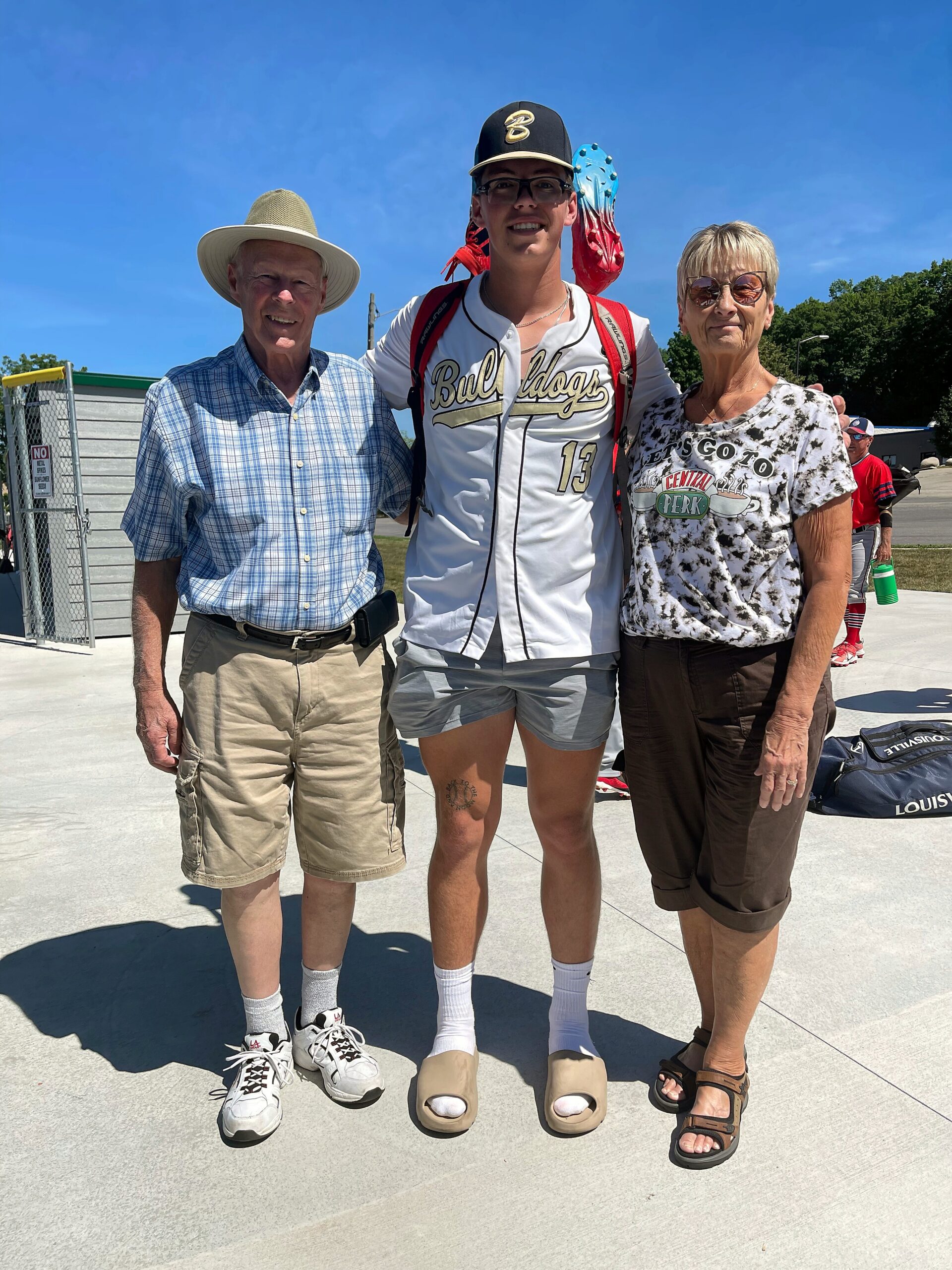 Three people posing outdoors on a sunny day: an older man in a straw hat, a baseball player in a white Bull Dogs jersey, and an older woman with sunglasses.