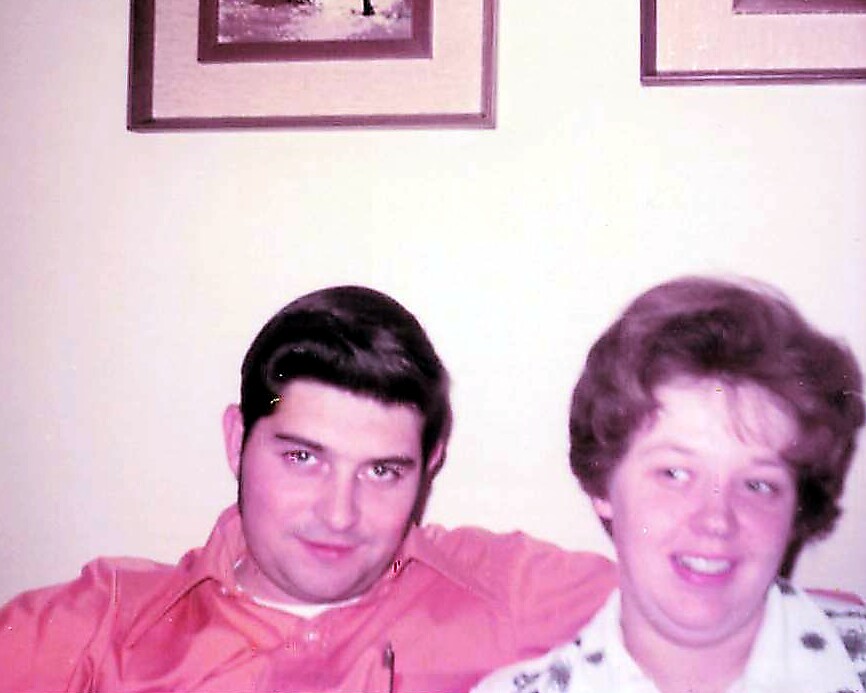 Young man with dark hair and a woman with short light hair sit close together on a couch, smiling at the camera.