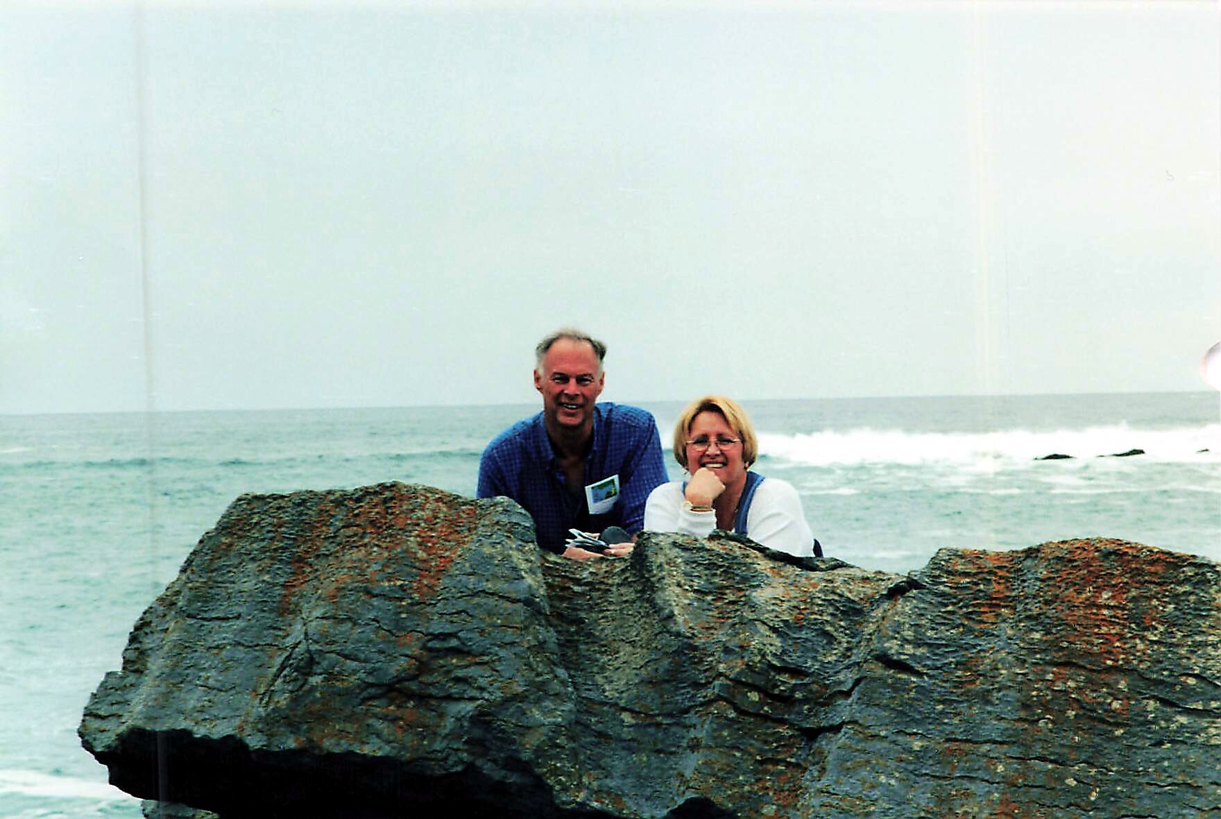 Man and woman sit on large rocks by the sea, smiling at the camera with waves behind them.
