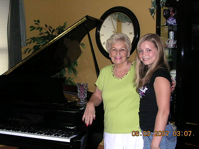 Two women stand beside a grand piano in a living room; an older woman in lime green top and a younger woman in black smile at the camera.