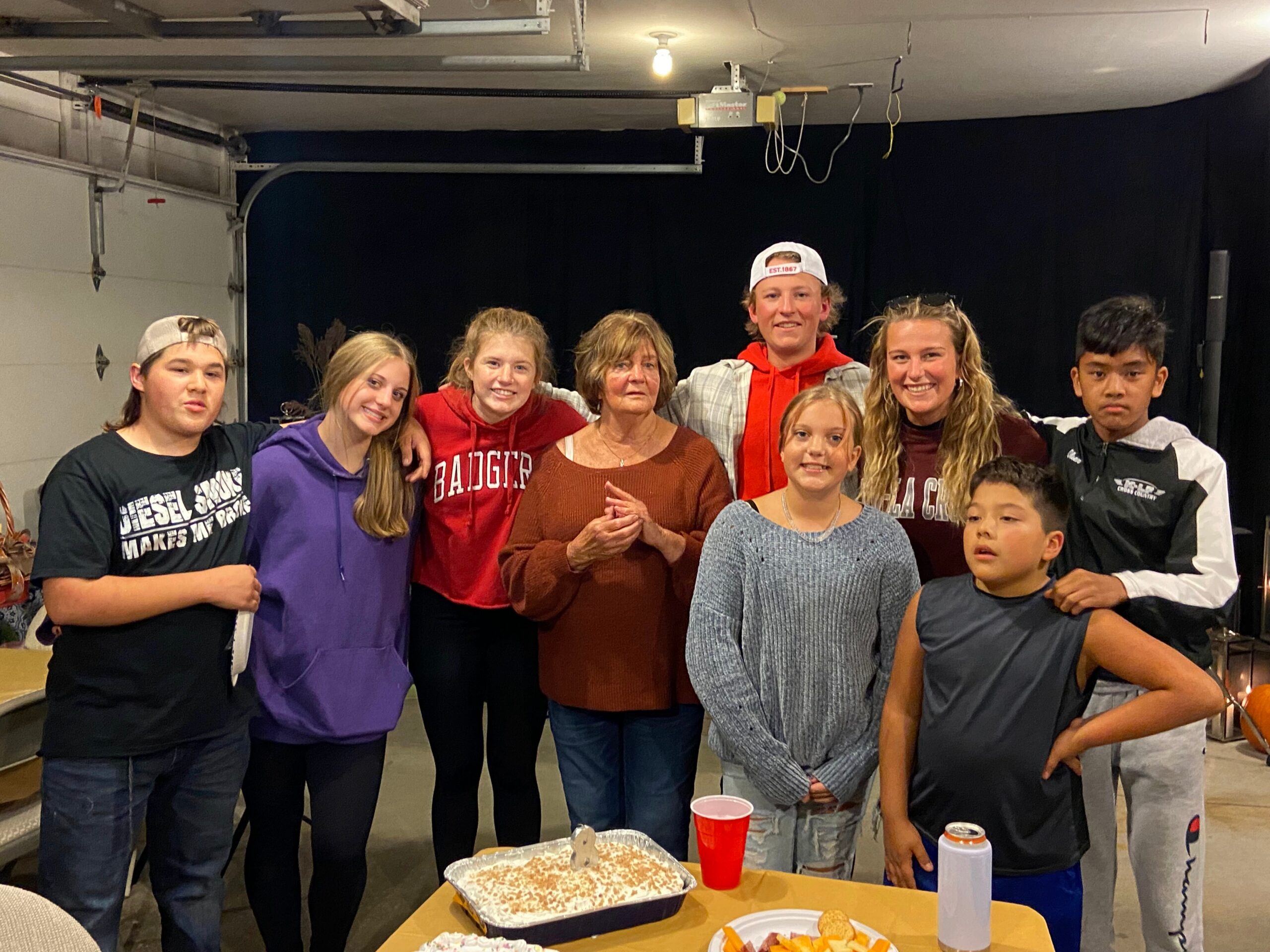 Group of people of various ages posing together in a garage-like space, with a cake and snacks on a table in front.