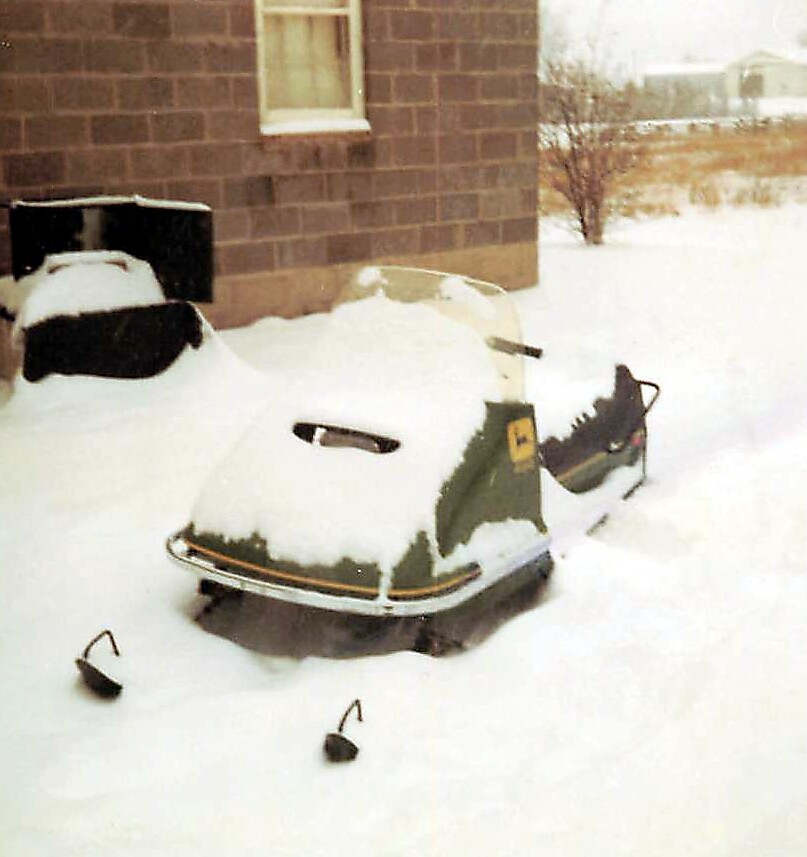 Snow-covered vintage snowmobile parked outside a brick house, with a thick layer of snow on its body and surrounding ground feel wintery.