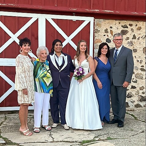 Group of six people posing outside a rustic venue: bride in white gown holding a bouquet, two men in suits, and three women in colorful dresses, standing in front of a red barn door and stone wall.