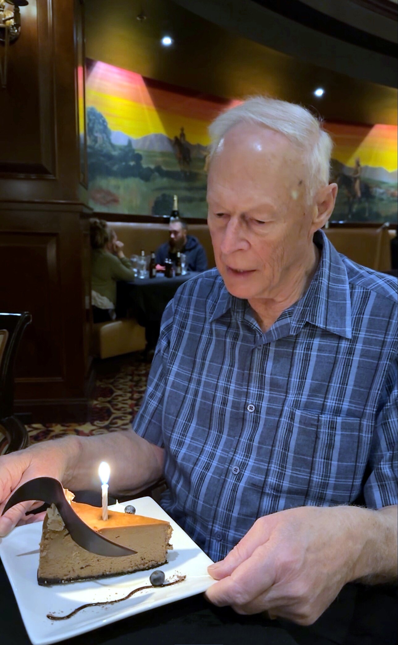 Older man in a blue plaid shirt at a restaurant table with a slice of chocolate cake and a lit candle in front of him, celebrating a birthday