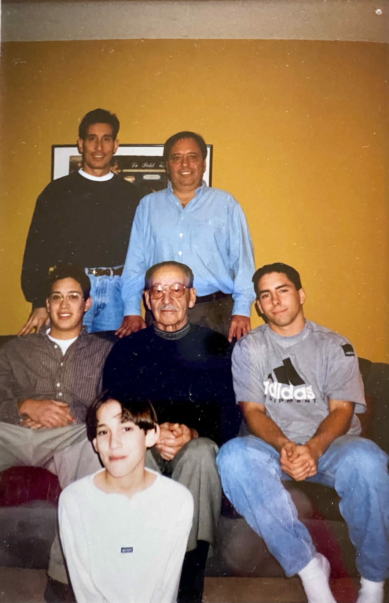 A family group photo in a living room with a yellow wall, five generations gathered together and smiling at the camera.