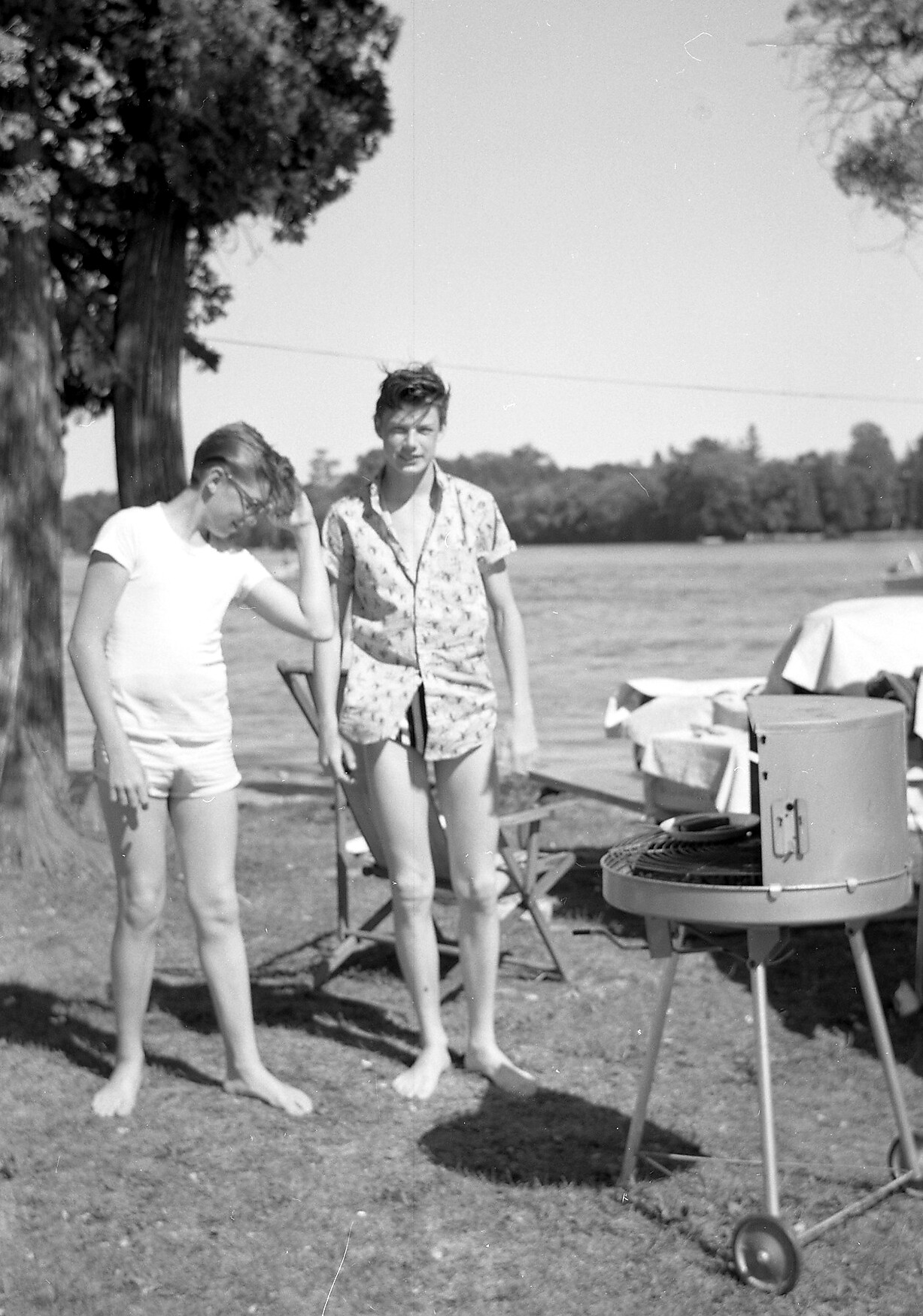 Two teens in swimwear stand by a round outdoor grill at a lakeside park, with tables and chairs in the background.