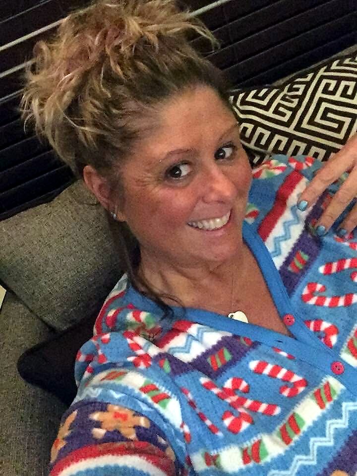 Smiling woman with a messy bun, wearing a bright candy-cane patterned blue sweater, relaxing on a gray couch.