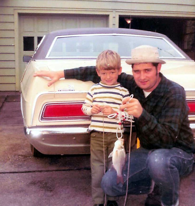 Father and young son posing by a beige vintage car, holding fishing lines with fish