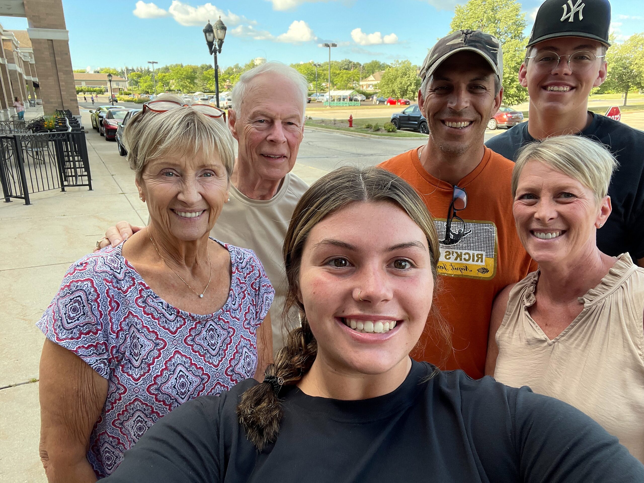 Group selfie of six smiling people outdoors in a parking lot with cars, trees, and a blue sky behind.