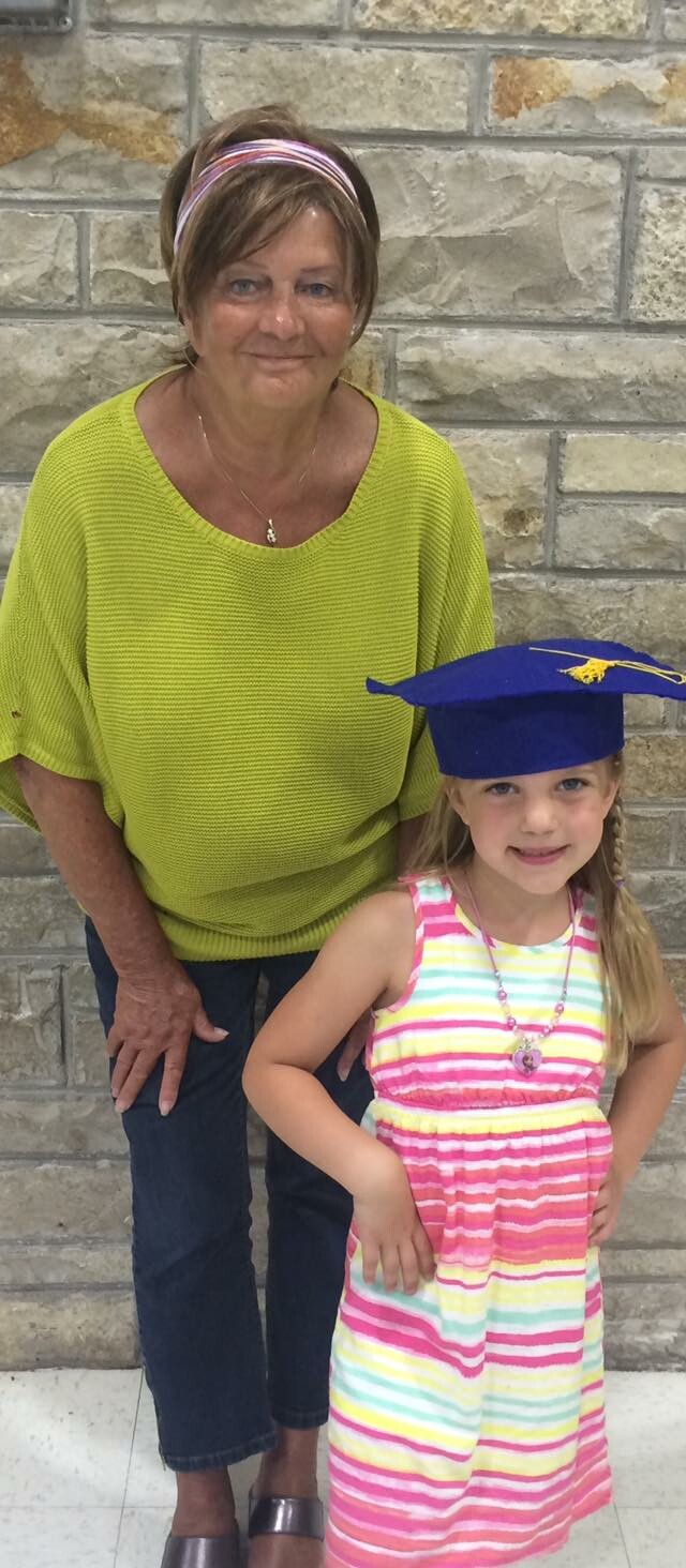 Older woman in a lime-green sweater stands beside a smiling young girl in a striped dress wearing a blue graduation cap, against a stone wall background.