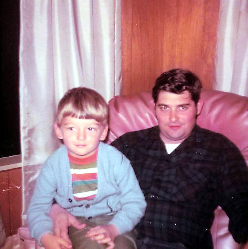 A man with dark hair sits beside a young boy on a pink armchair in a cozy living room with wood paneling and curtains.