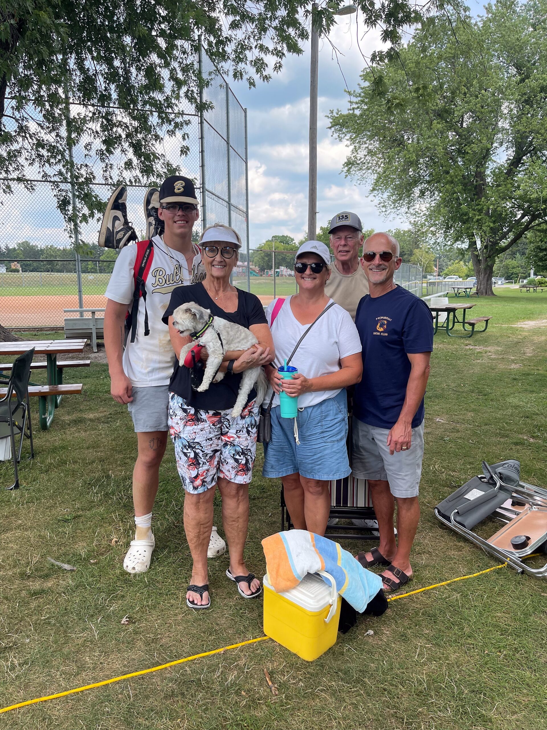 Group of five adults outdoors at a baseball field, holding a small white dog and smiling near gear and a yellow cooler.