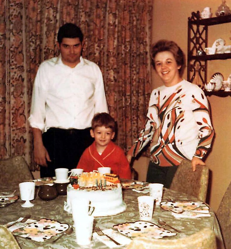Three people stand around a birthday cake on a dining table: a smiling woman on the right, a man on the left, and a young boy in a red outfit at the center front.