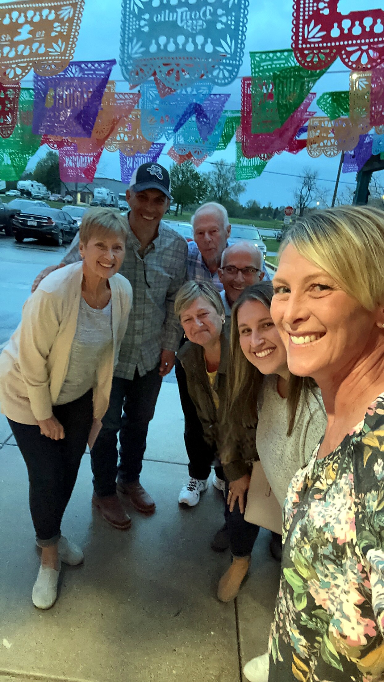 Smiling group of six adults posing for a selfie under colorful decorative paper banners outdoors.