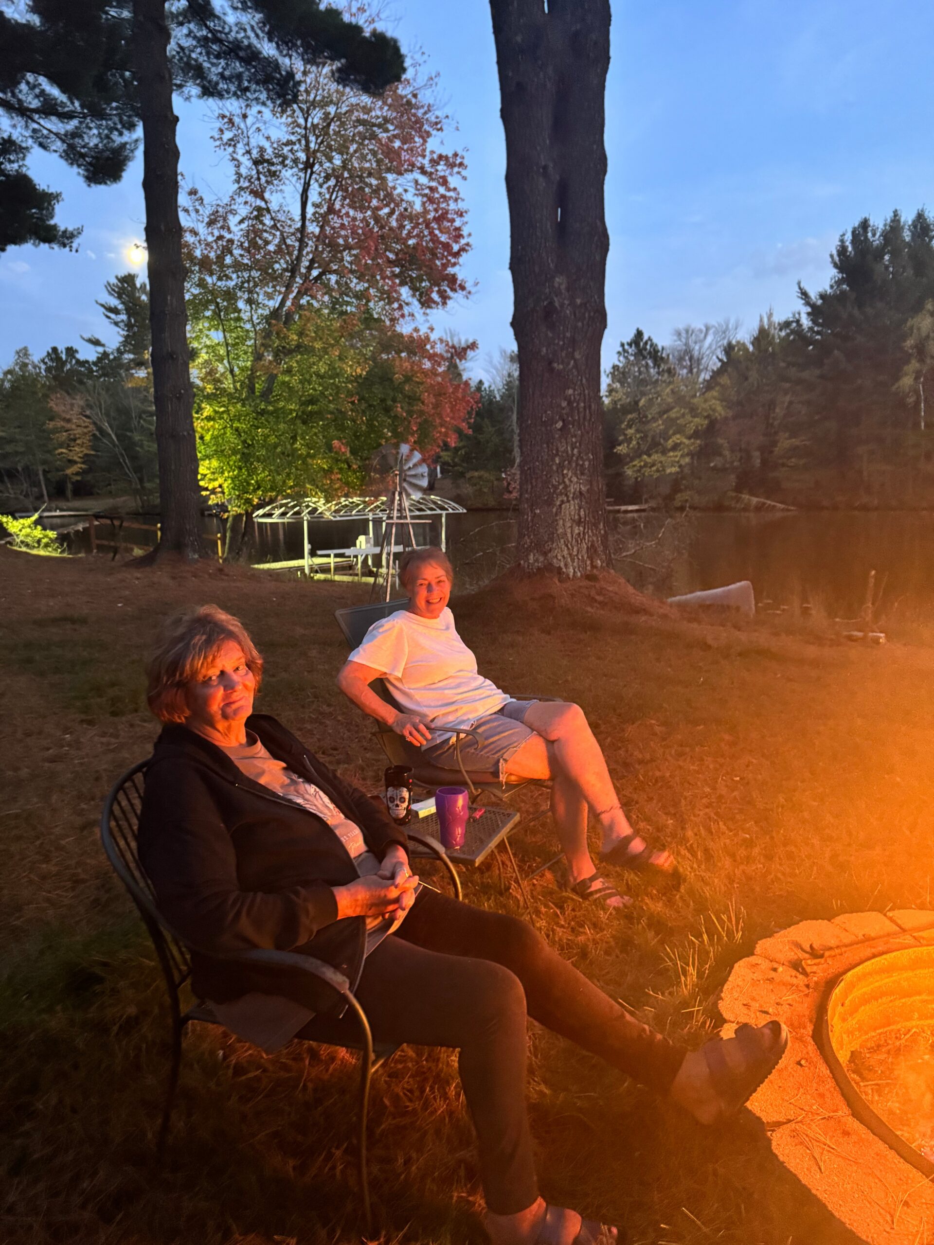 Two people sit in lawn chairs by a campfire at dusk, with trees and a lake in the background and warm firelight on their faces.