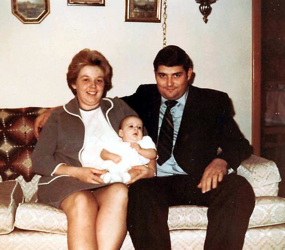 A couple sits on a patterned sofa holding a baby in a living room setting with framed walls decor behind them.