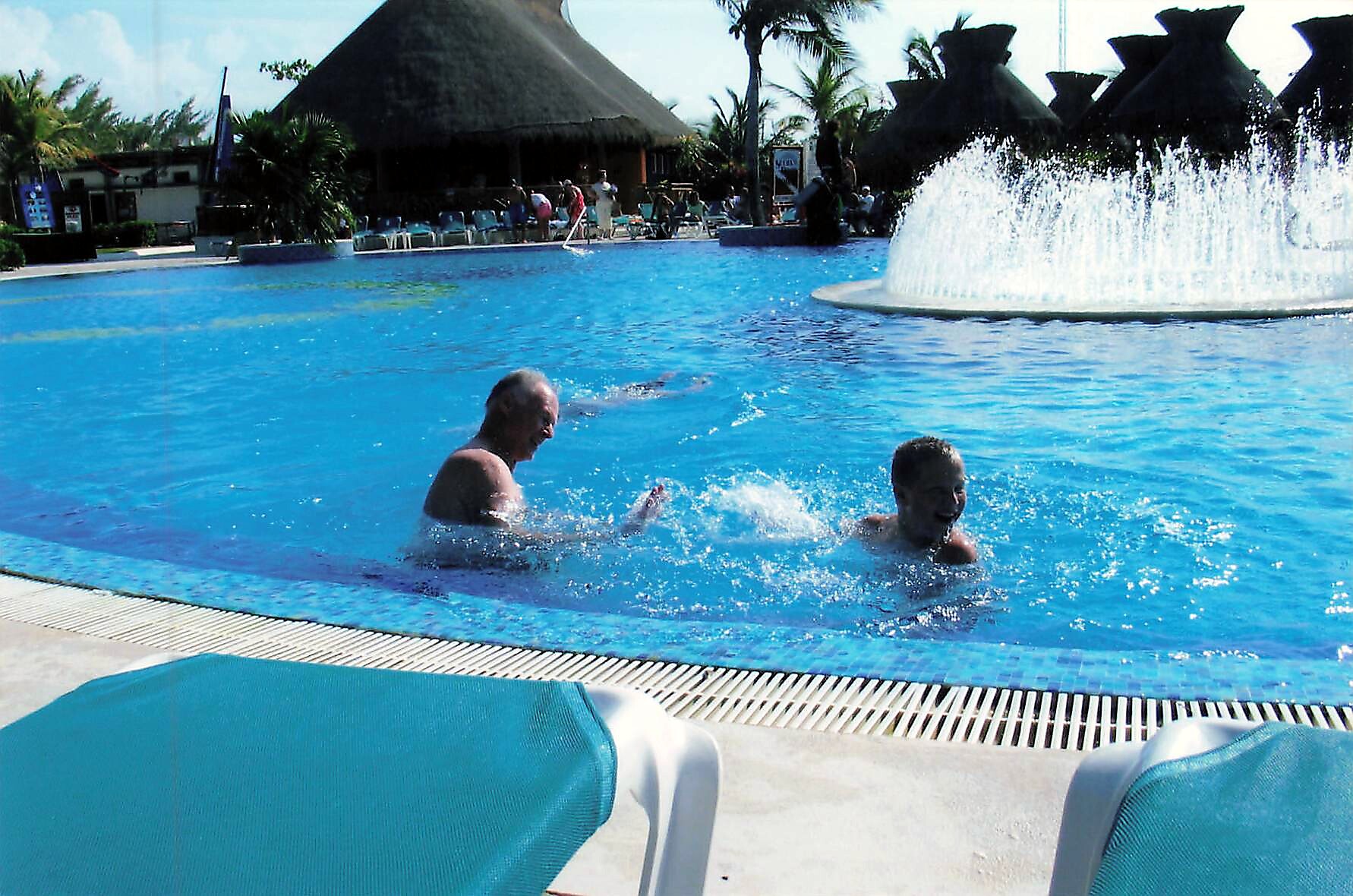 Two men swim and play in a bright blue pool, with a large fountain and thatched-roof huts in the background.