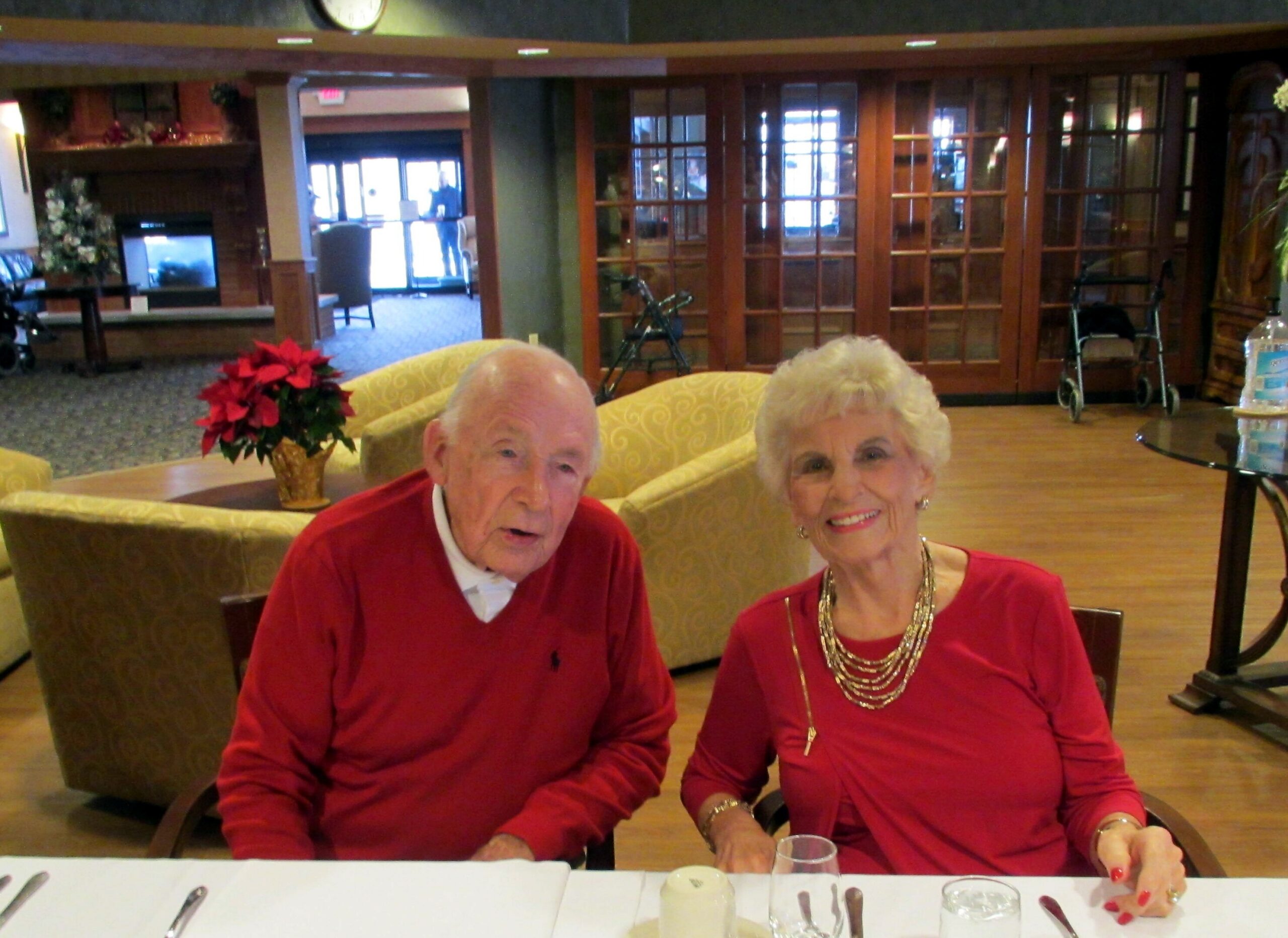 Older couple in red sweaters sit at a dining table in a holiday lobby, smiling with utensils and glasses on the table in front of them.