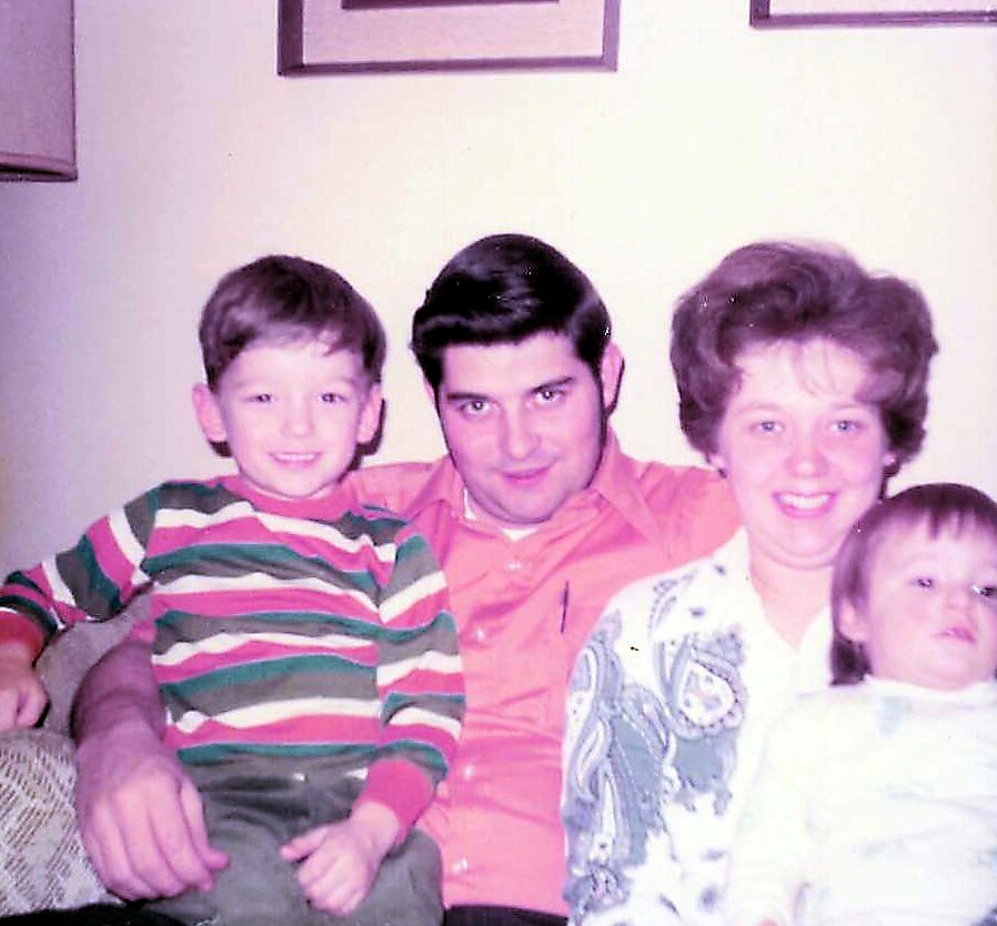 Family of four sitting close together on a couch: father in pink shirt, mother in patterned blouse, older boy in a striped sweater, and a baby girl beside them.