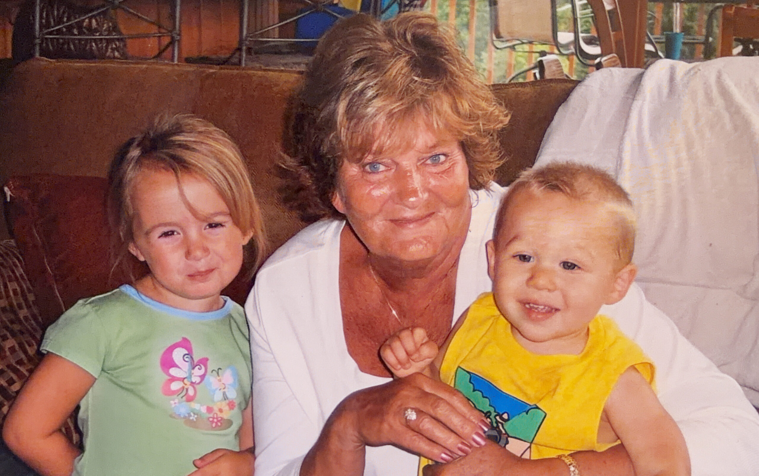 A smiling older woman sits on a couch with two young children, one on each side, in a cozy living room setting.