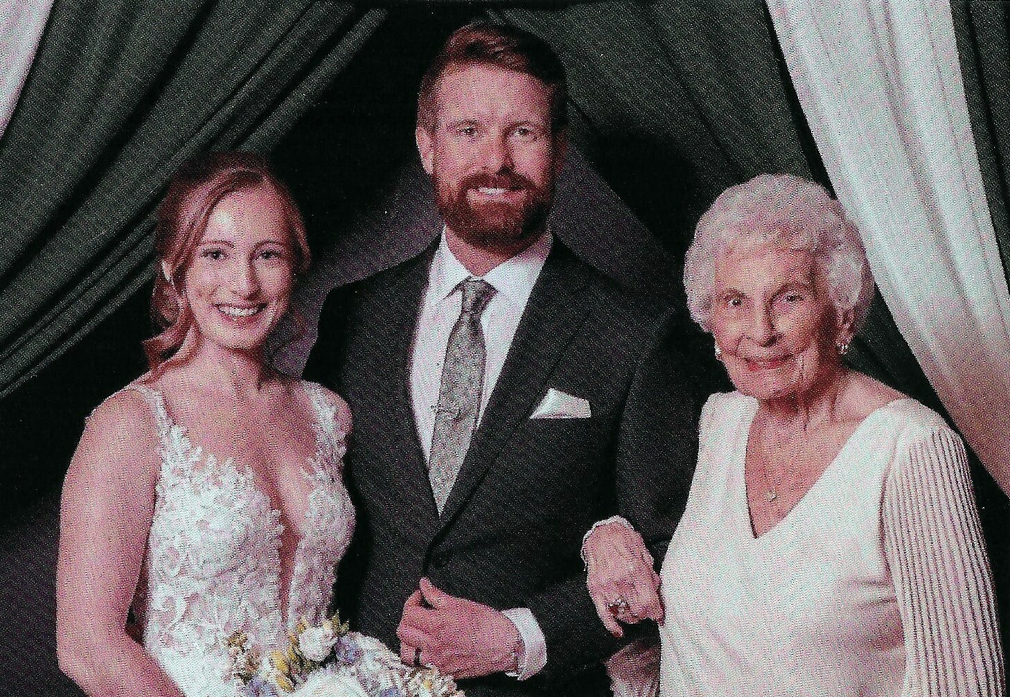 Bride in a lace white gown and bouquet, on the left, stands with a bearded groom in a dark suit, flanked by an older woman in white on the right in a wedding setting.
