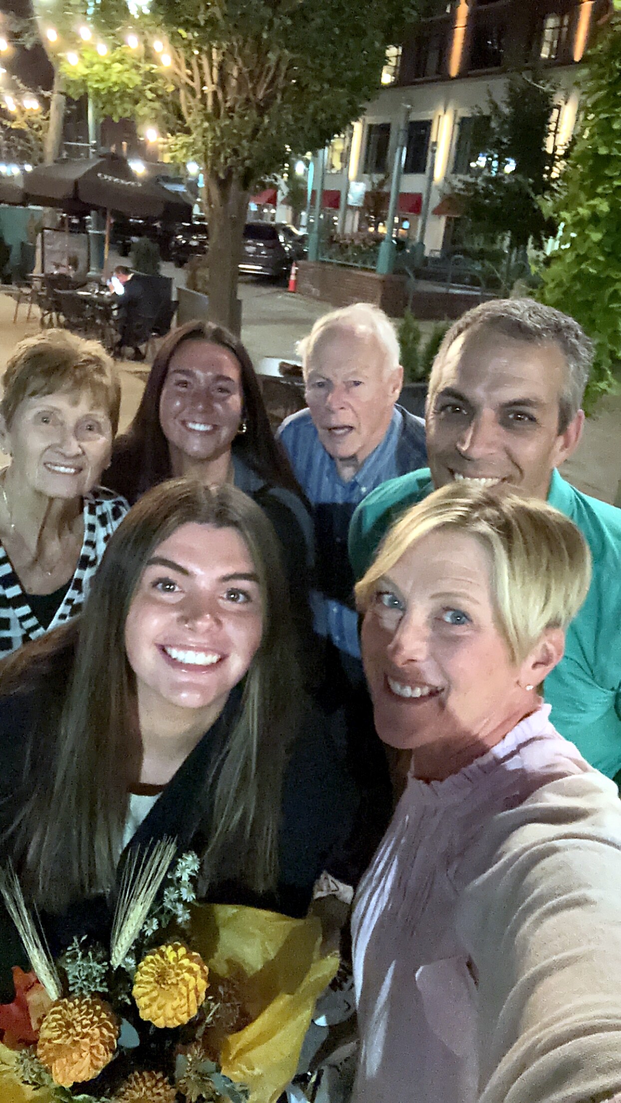 Group of six people smiling for a selfie on a city sidewalk at night, with a bouquet of flowers in the foreground