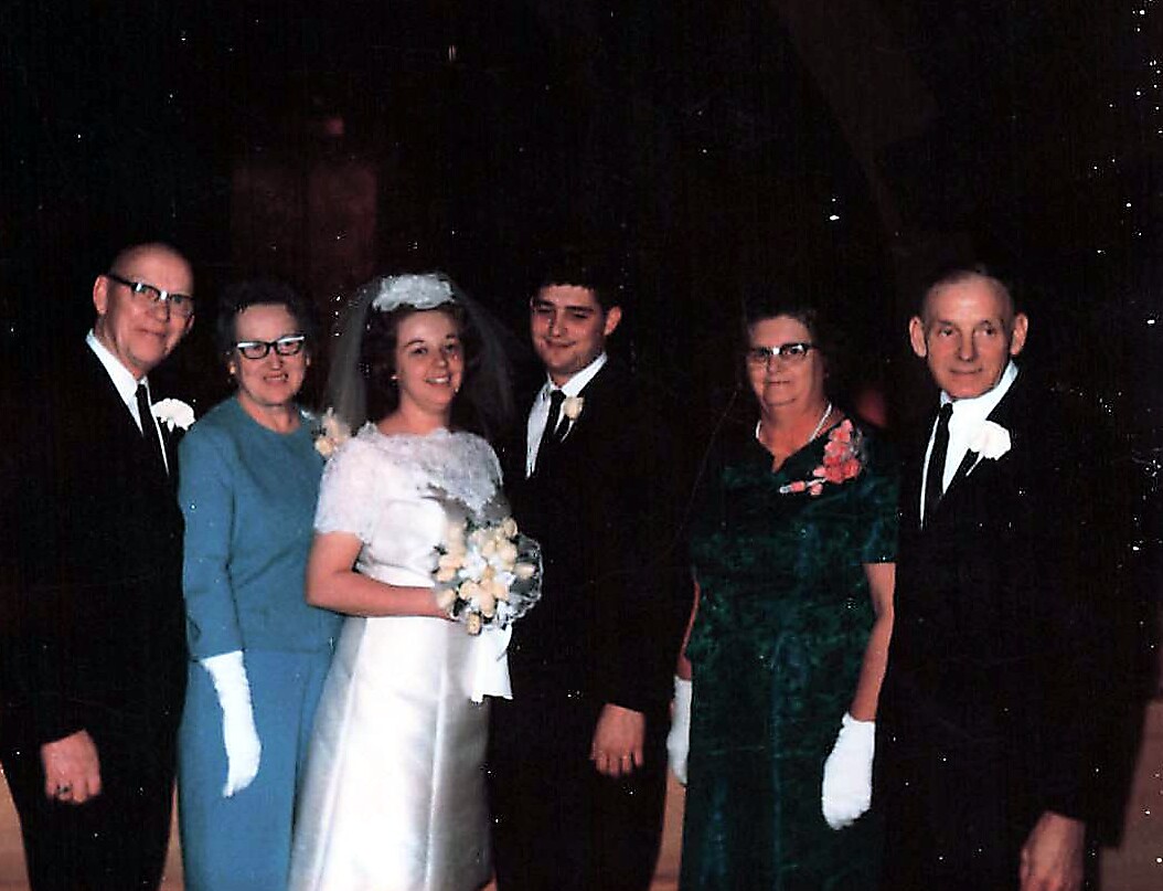 Bride in white gown holding bouquet, standing with groom and family in formal attire at a wedding reception scene.
