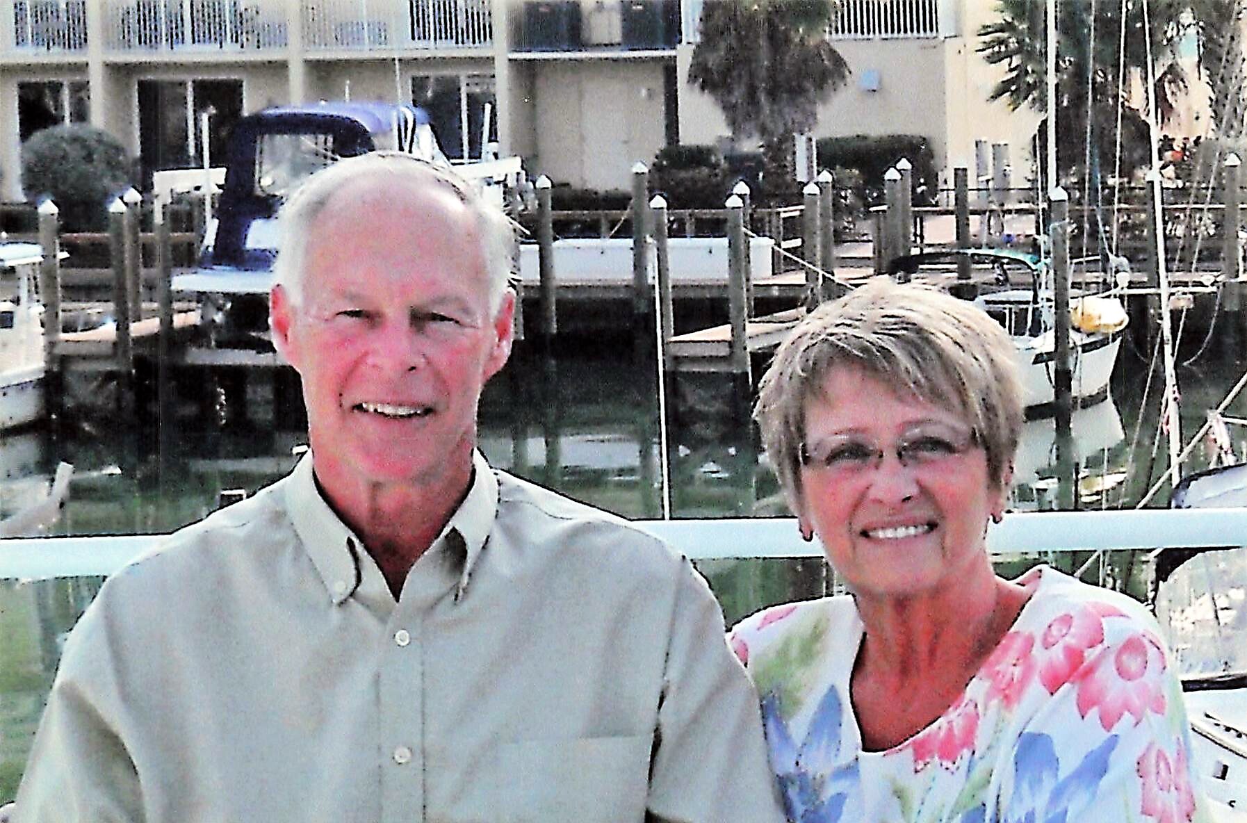 Smiling elderly couple posing at a marina, with boats and dock in the background.