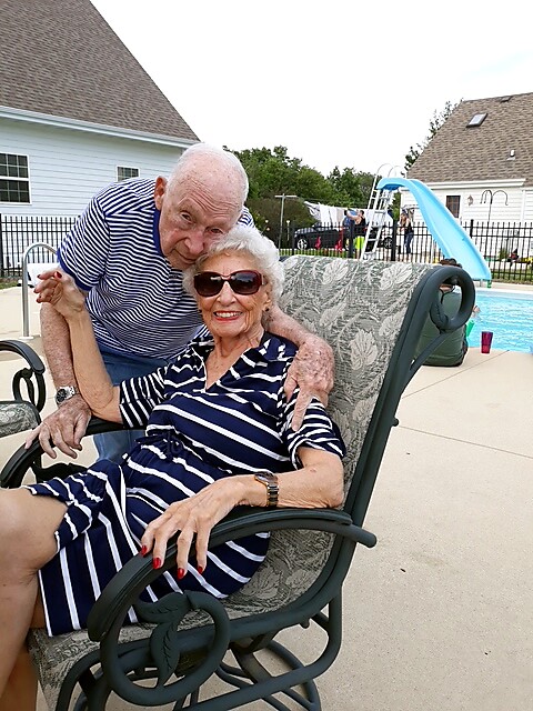 Older couple at a sunny backyard pool; man stands behind, holding the seated woman in a striped dress, both smiling.