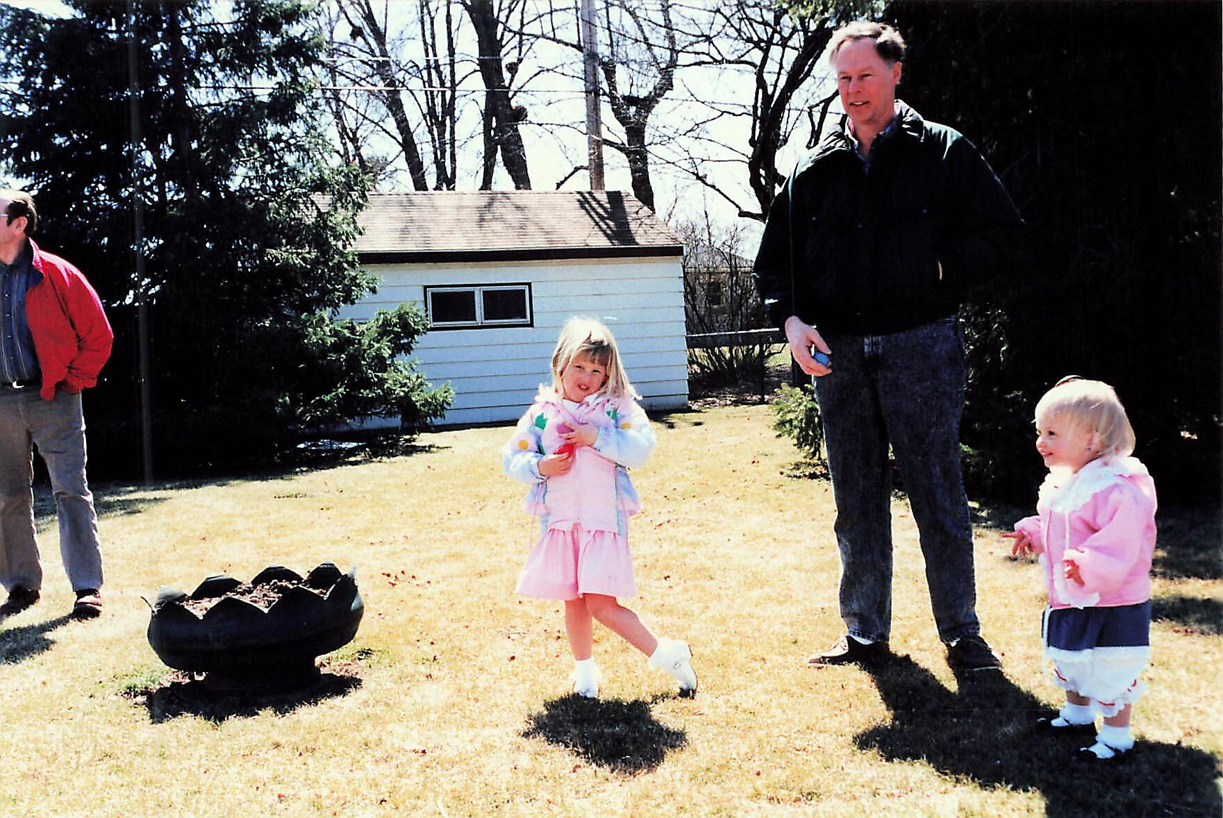 A man in a red jacket stands to the left while two young girls in pink jackets play on a sunny yard near a white shed and a tire obstacle in the foreground.