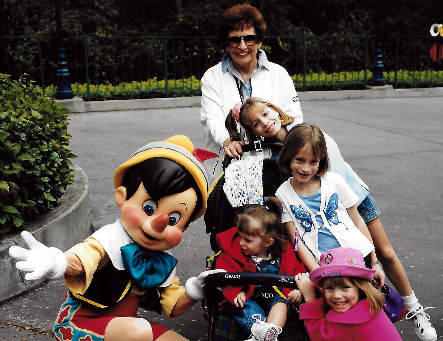 Costumed character wearing a yellow hat leans toward smiling children as they ride in a stroller, with an adult woman in a white jacket standing behind.