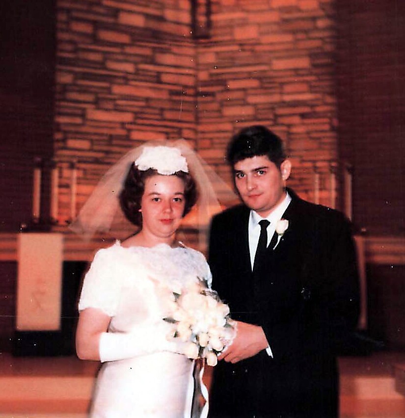 Bride in a white wedding gown and veil stands beside a groom in a dark suit, holding a bouquet in a church setting (vintage photo)