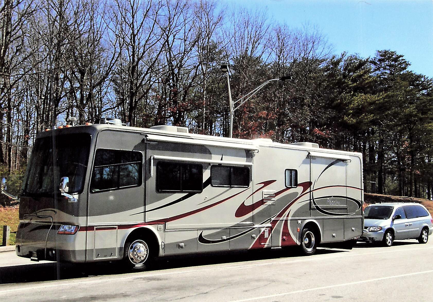 Large grey Class A motorhome with red and black swoosh graphics, parked in a lot with leafless trees in the background and a silver car behind it.