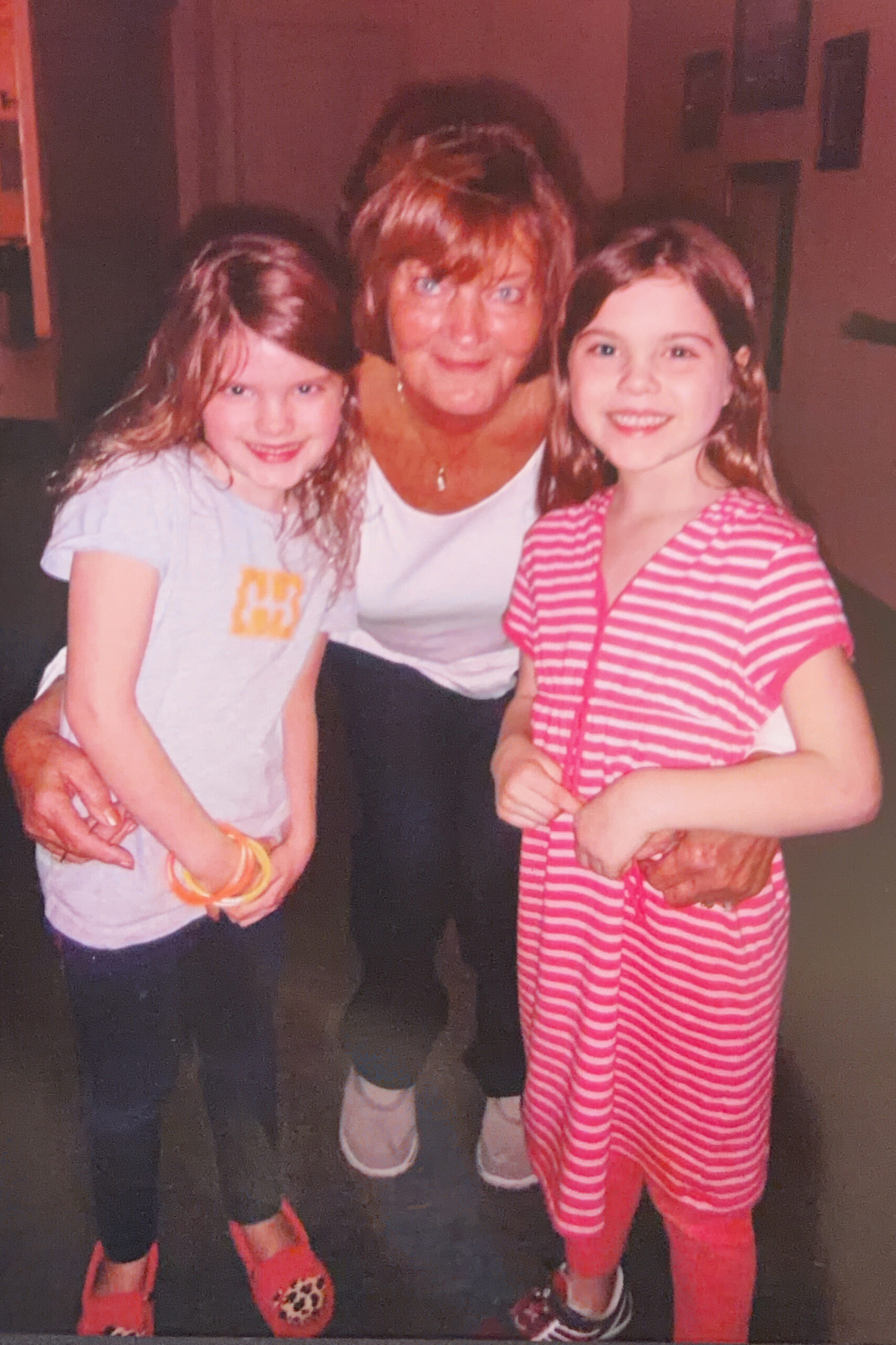 Three people posing together indoors: a woman in the center flanked by two smiling girls on either side.