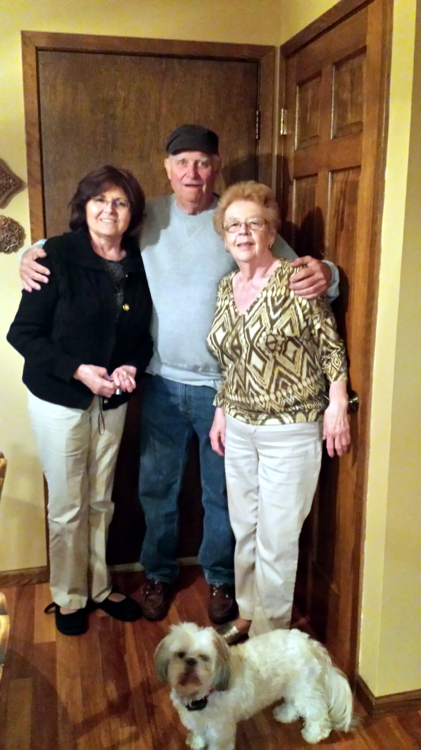 Three adults pose together in a doorway with two small fluffy dogs at their feet in the hall.