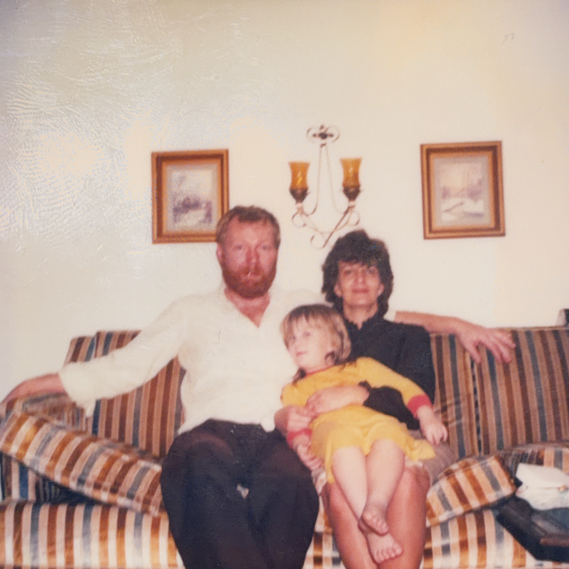 A family of four sits together on a striped sofa in a living room with framed pictures on the wall behind them.