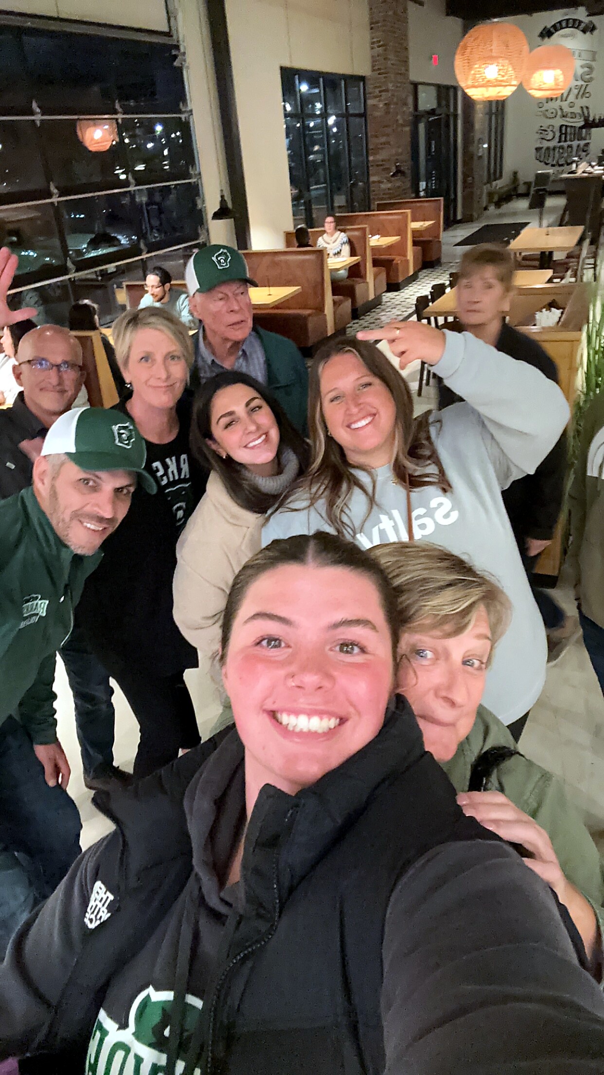 Group of people taking a selfie in a casual restaurant, all smiling at the camera with booths and warm lighting in the background.