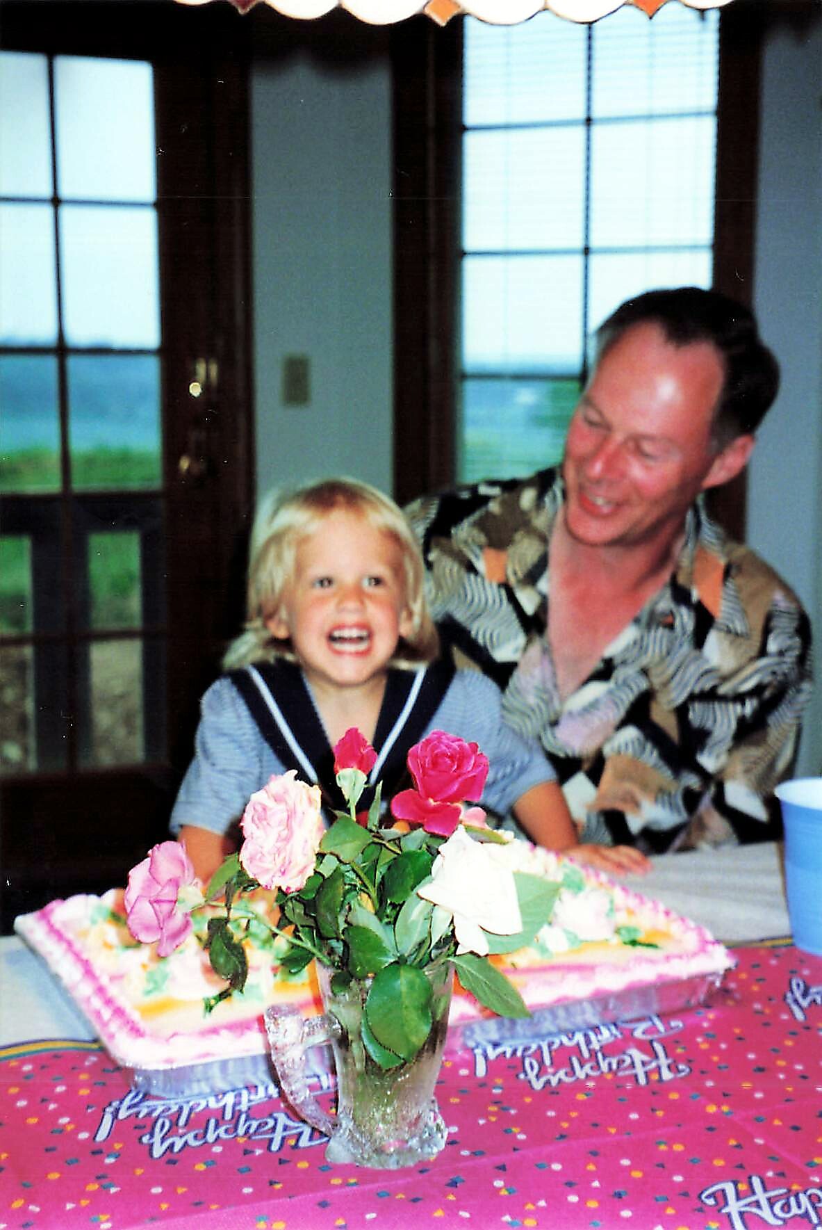 Young girl with blonde hair smiling and laughing at a birthday party, with a flower centerpiece and a man smiling beside her at a decorated table.
