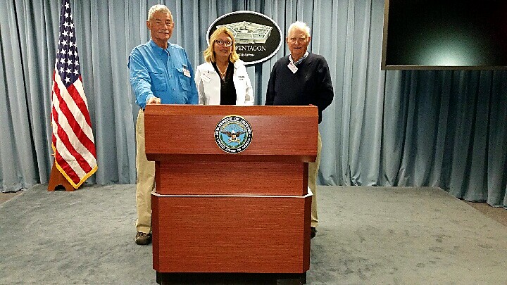 Three people stand behind a wooden podium with a government seal, US flag visible to the left in a briefing room.