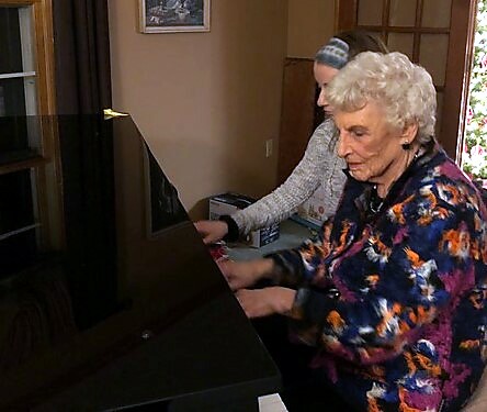 Two elderly women playing a black grand piano in a home setting; foreground woman with short white hair wearing a colorful floral jacket.