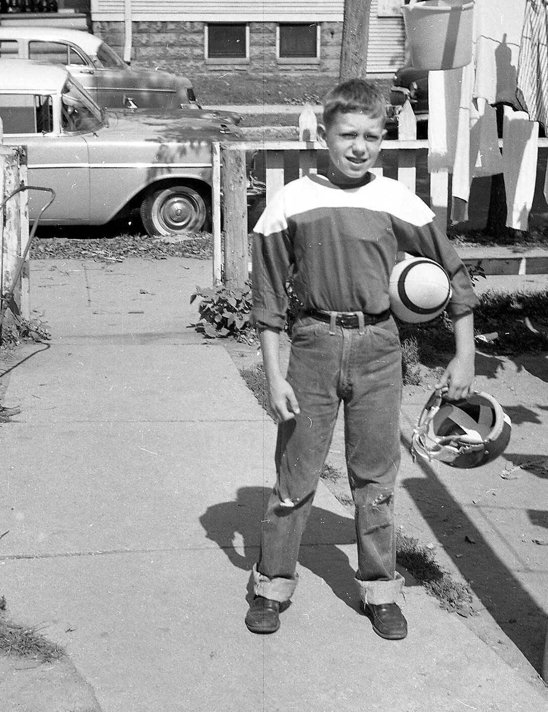 Boy standing on a sunny sidewalk, holding a ball under his arm and a helmet in his hand, wearing a striped shirt and rolled cuffs jeans with sneakers; cars and a clothesline backdrop.