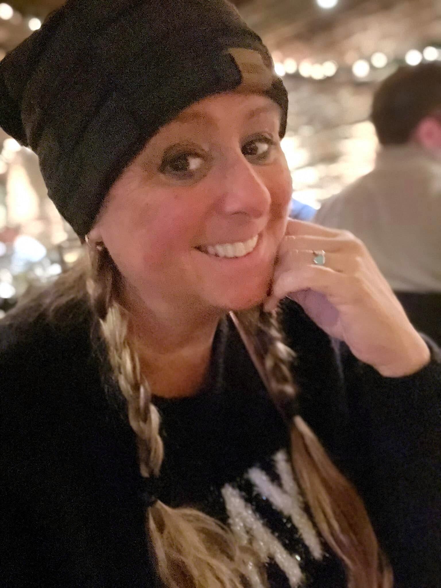 Close-up portrait of a smiling woman wearing a black beanie, with braided hair and a turquoise-ringed hand near her chin.